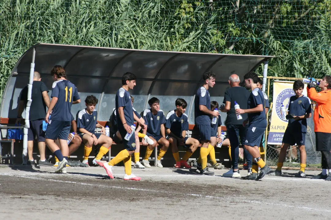 Giovani calciatori del Pian Due Torri in divisa blu e gialla seduti e in piedi vicino alla panchina durante un momento di pausa.